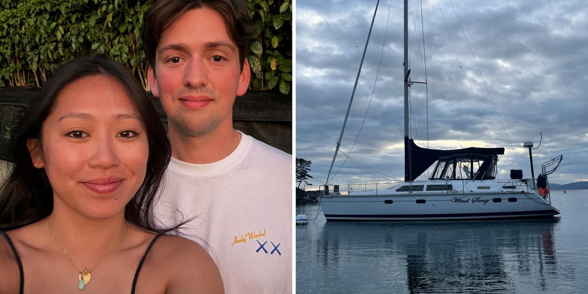 A couple standing on the deck of a sailboat, smiling, with a scenic water view behind them.