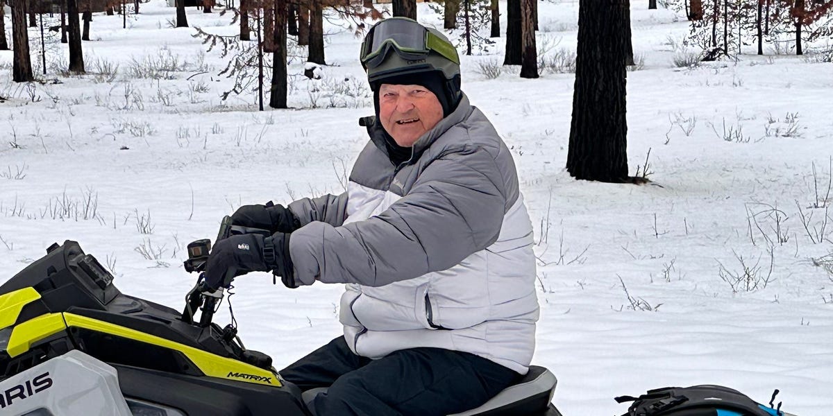 John Adams, an 80-year-old engineer, is shown smiling outdoors, wearing a pilot's headset and jacket.