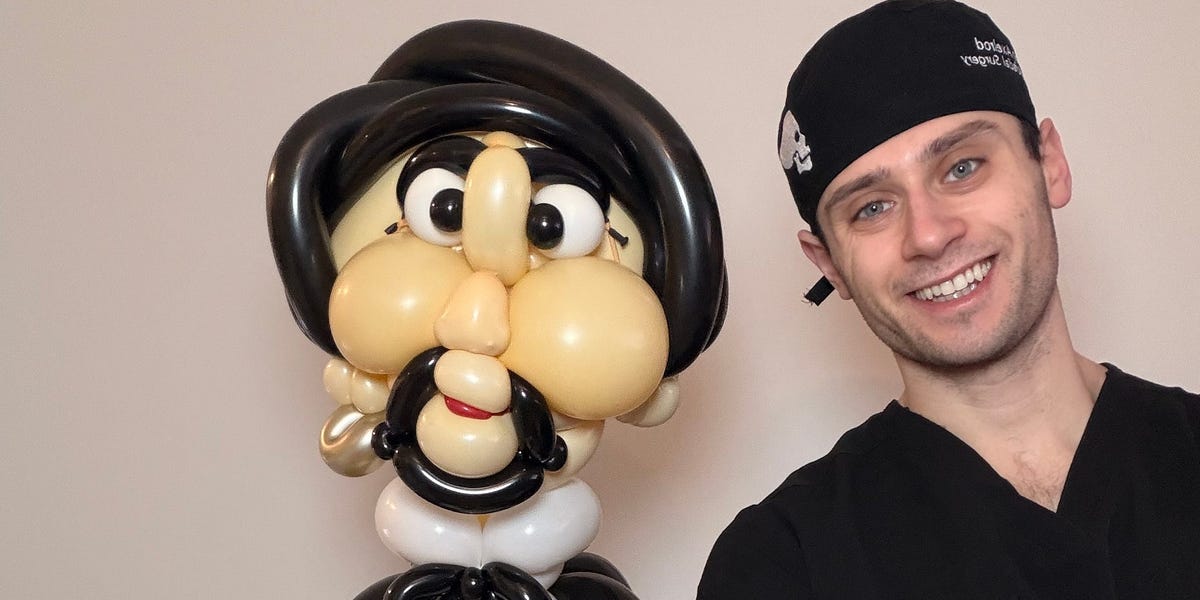 A young man in scrubs smiling and holding a complex balloon sculpture.