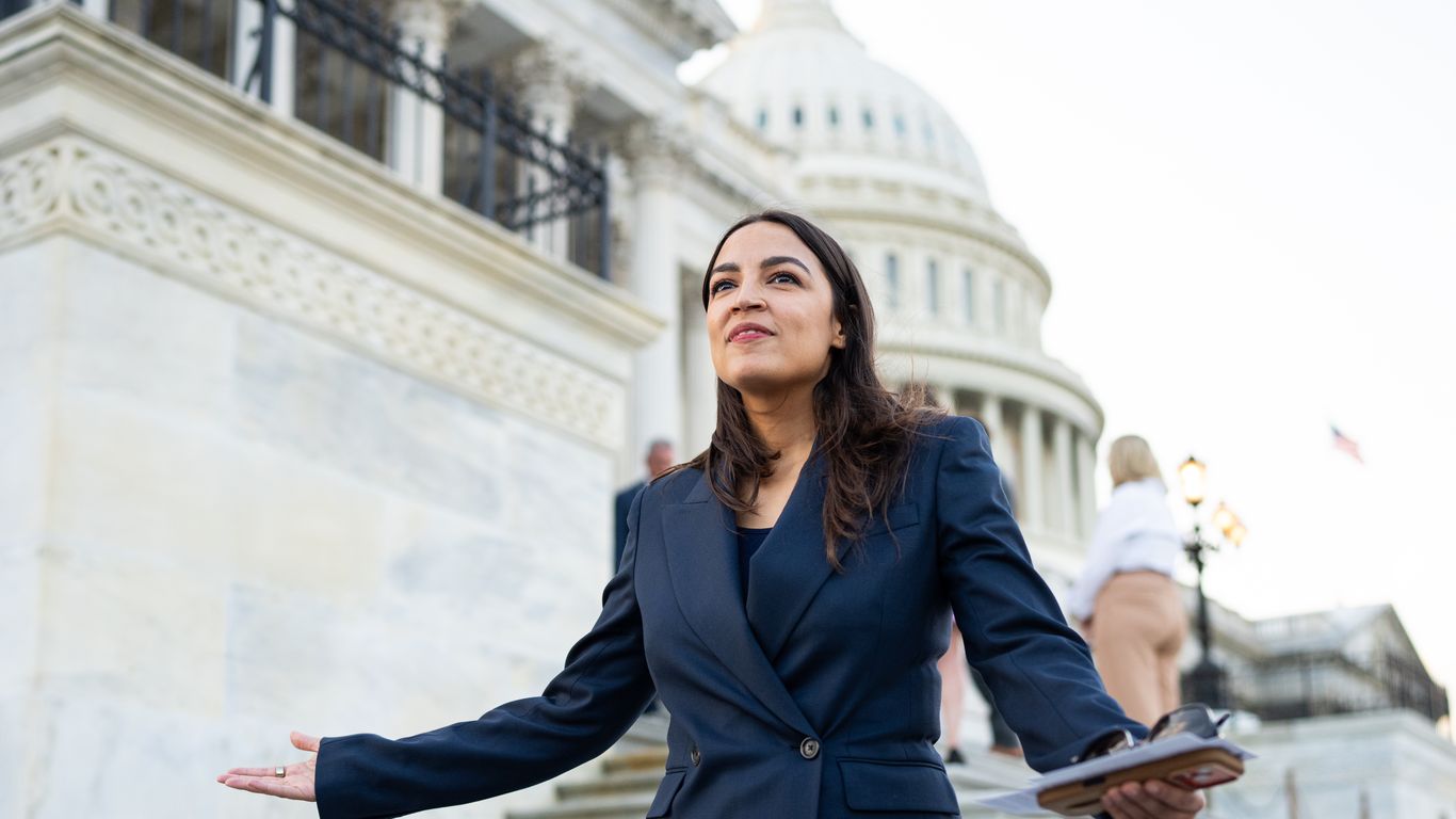 Rep. Alexandria Ocasio-Cortez speaking at a podium with a blurred press corps in the background.